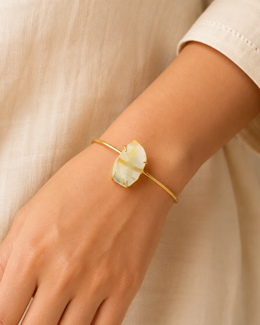 A gold bangle bracelet with a large, organic-shaped cream-colored stone, shown worn on a person's wrist against a soft beige linen background.