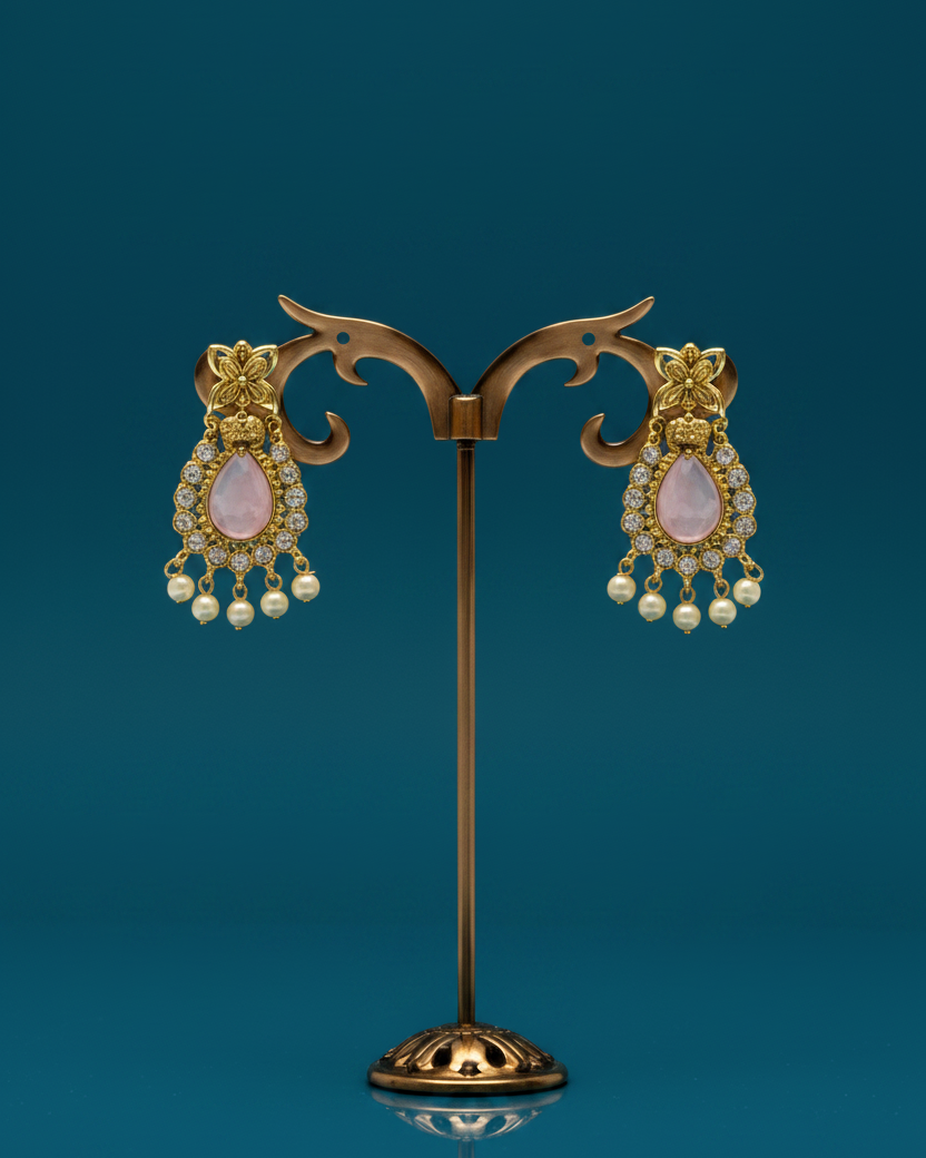 A pair of ornate gold-toned dangle earrings with pale pink teardrop-shaped stones and rows of small white pearls, displayed on a decorative stand against a dark teal background.
