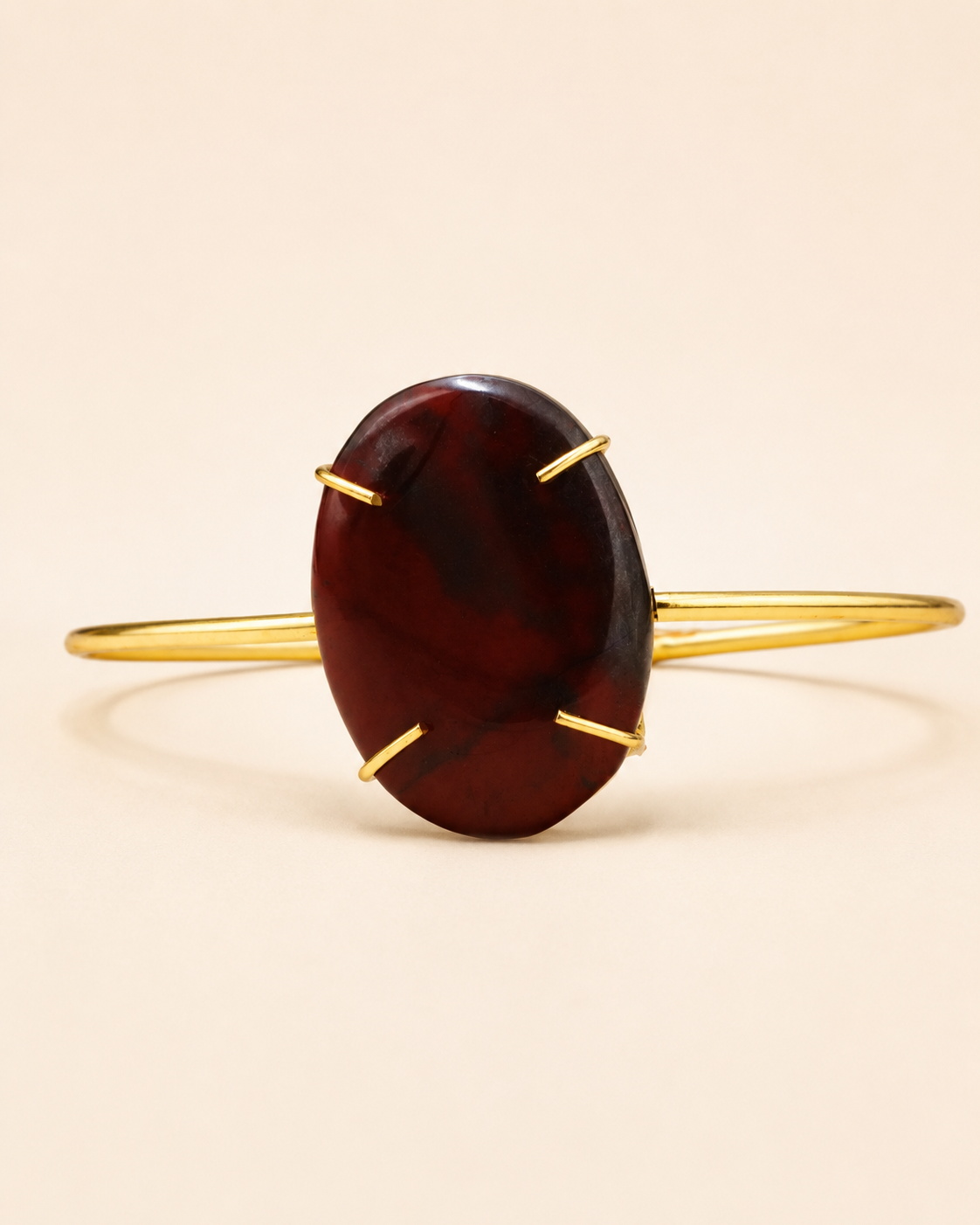 A gold-plated cuff bracelet featuring a large, polished oval cabochon stone in a deep burgundy red color, held in place by four prongs, set against a solid light beige background.