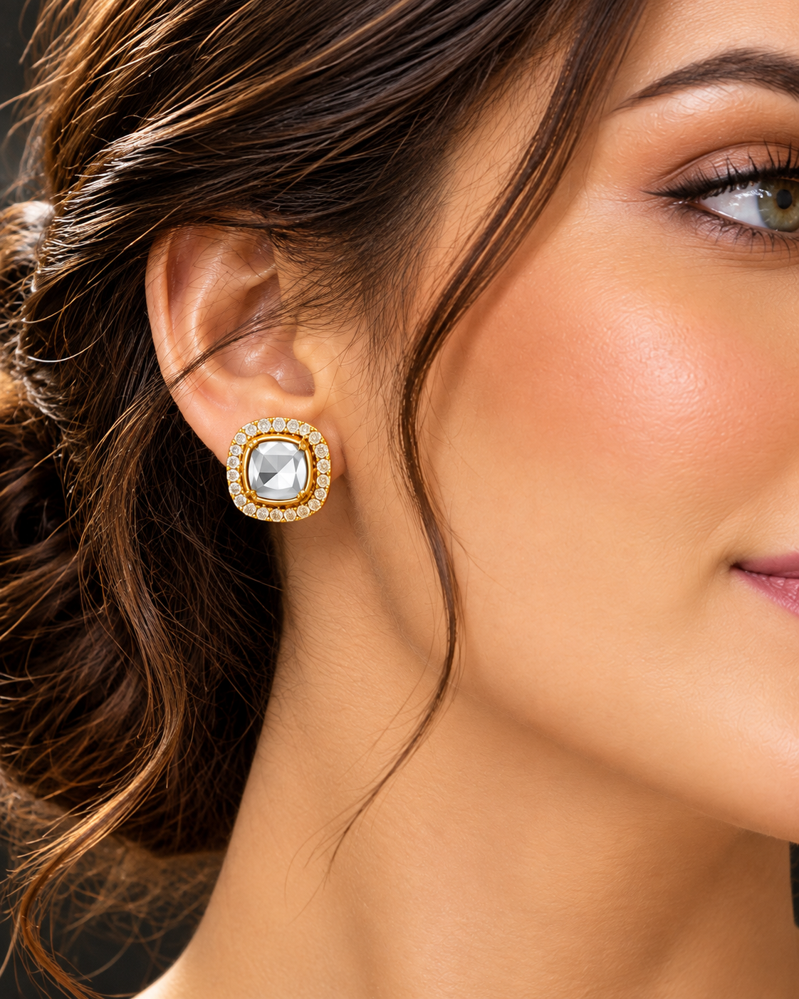 A close-up of a woman's ear wearing a square gold-toned earring with a clear central stone and a border of small clear stones.
