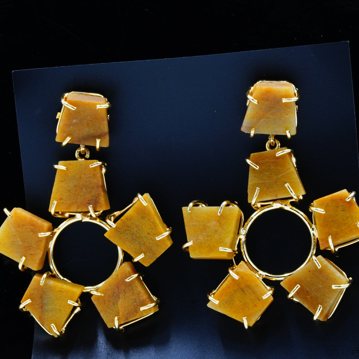 A pair of chunky gold-tone statement earrings featuring multiple irregular amber-yellow stone clusters arranged around a central open circle, displayed on a dark backing card against a black background.