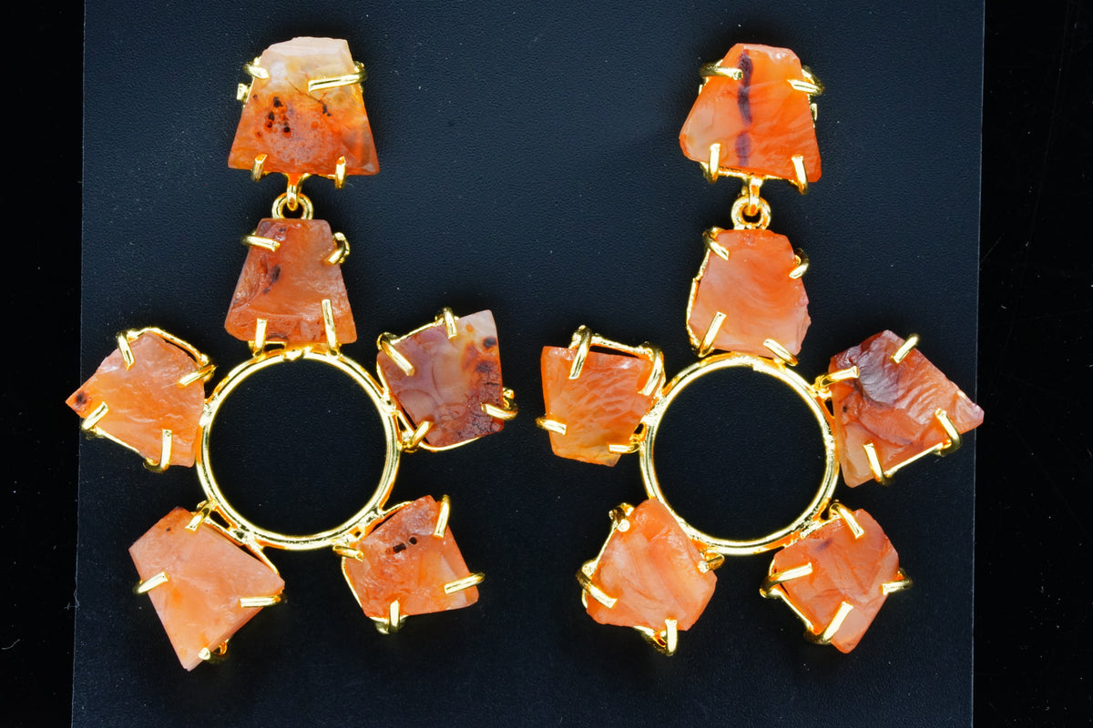 A pair of large statement dangle earrings featuring irregular orange stone pieces in gold-toned settings, arranged around a central gold ring on a black background.