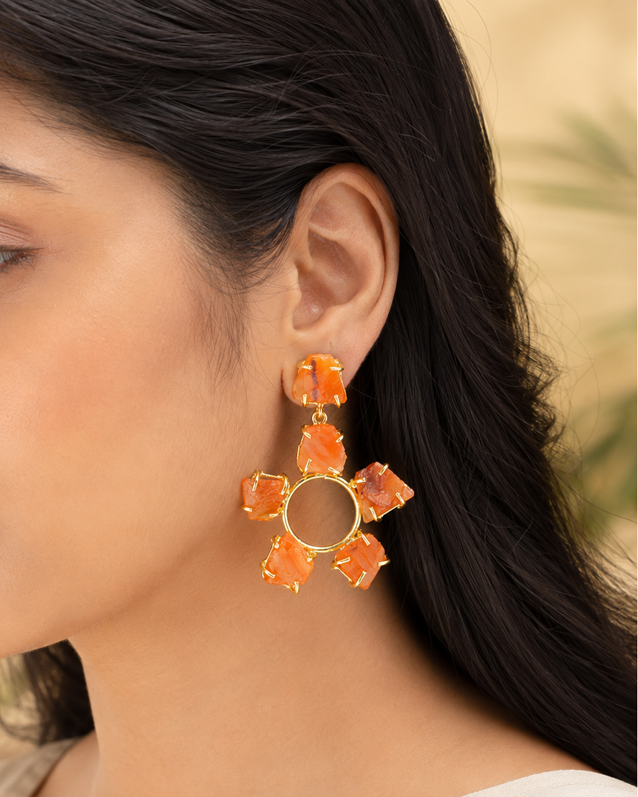 A close-up of a woman wearing a large gold-toned dangle earring with six orange stone-like petals arranged in a flower shape.