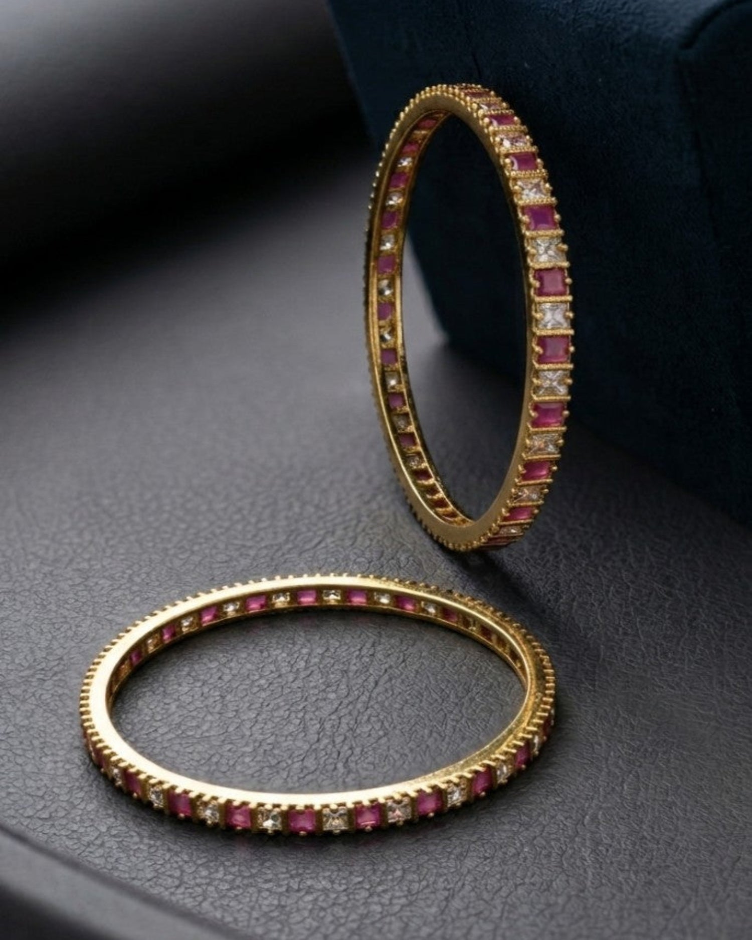 Two gold bangles with red and clear gemstones on a dark surface