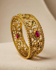 An ornate gold filigree bangle bracelet with inlaid pinkish-red and green gemstones, displayed on a textured beige surface.