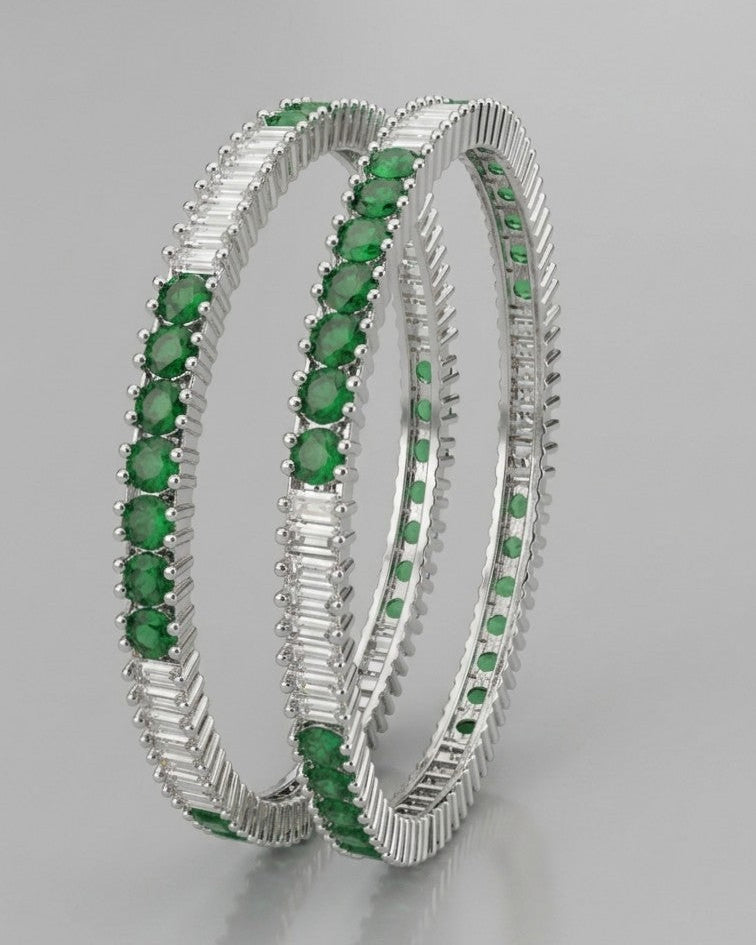 Pair of rhodium-plated silver bangles featuring a row of round emerald green CZ stones bordered by baguette-cut clear stones.
