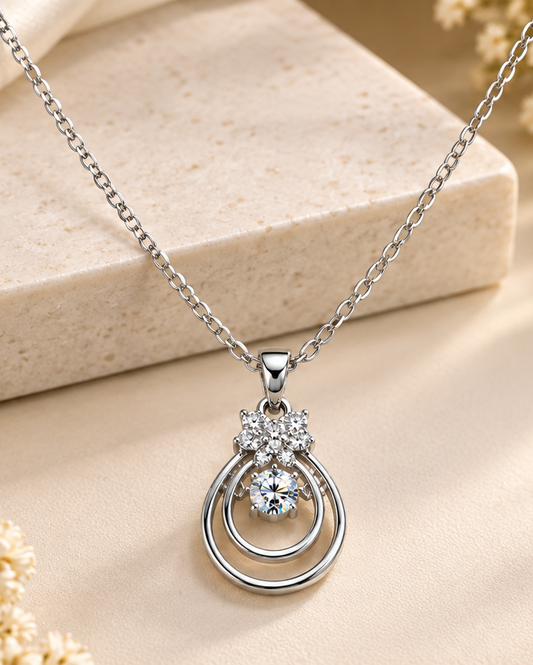 A silver-tone pendant necklace with a double teardrop ring design and a small floral cluster of clear stones, displayed on a beige textured surface with a stone block.