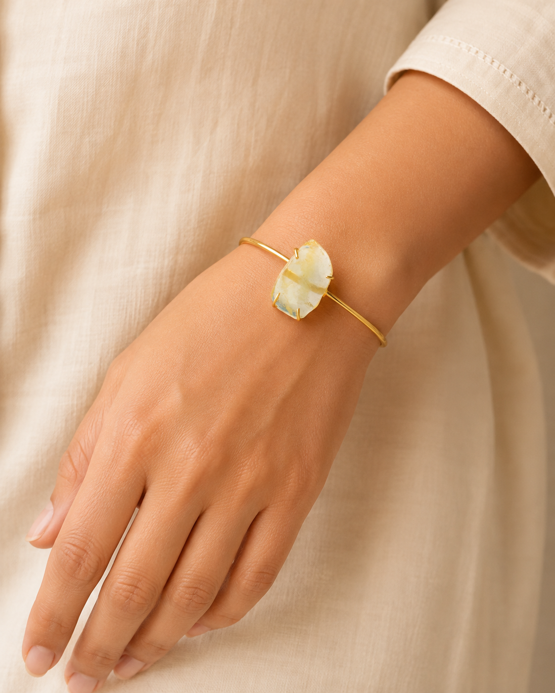 A gold bangle bracelet with a large, organic-shaped cream-colored stone, shown worn on a person's wrist against a soft beige linen background.