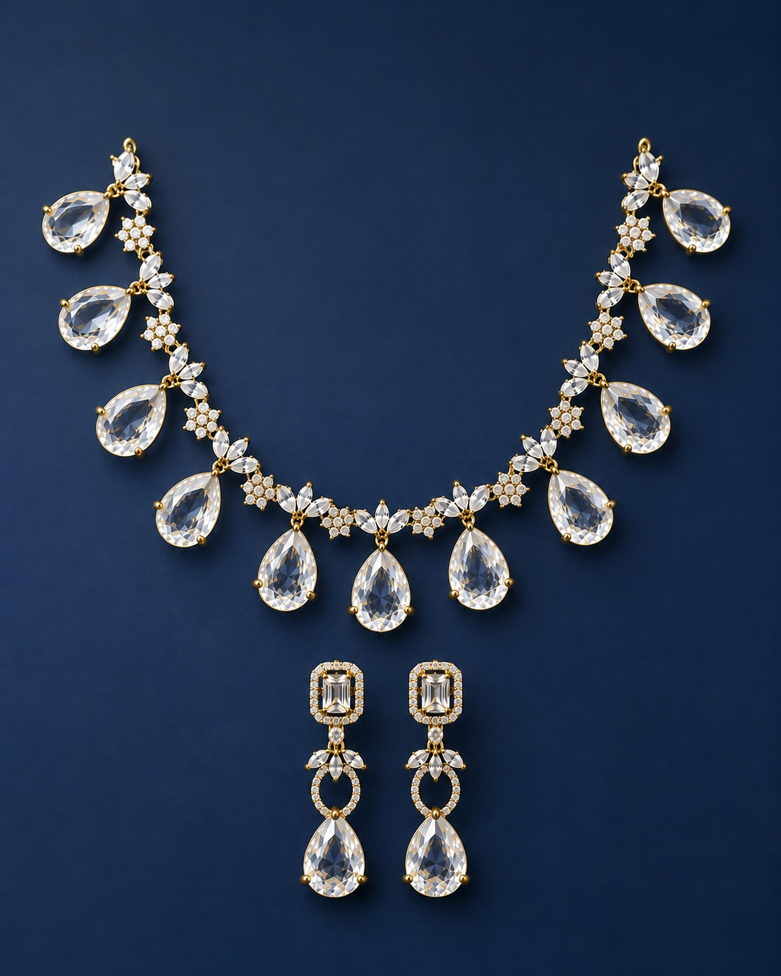 A gold-tone jewelry set featuring a necklace and matching drop earrings, both adorned with clear teardrop-shaped gemstones and small floral star motifs, displayed on a dark navy blue background.