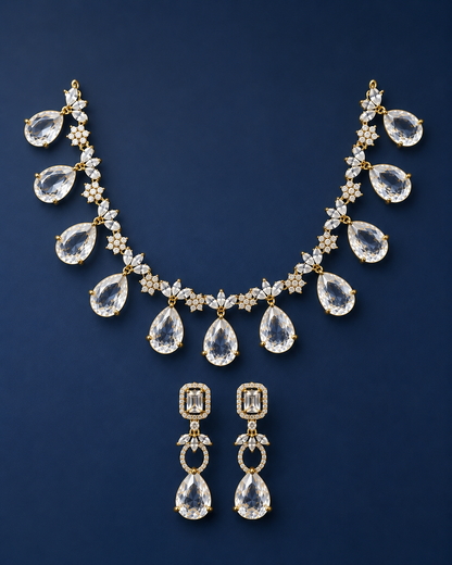 A gold-tone jewelry set featuring a necklace and matching drop earrings, both adorned with clear teardrop-shaped gemstones and small floral star motifs, displayed on a dark navy blue background.