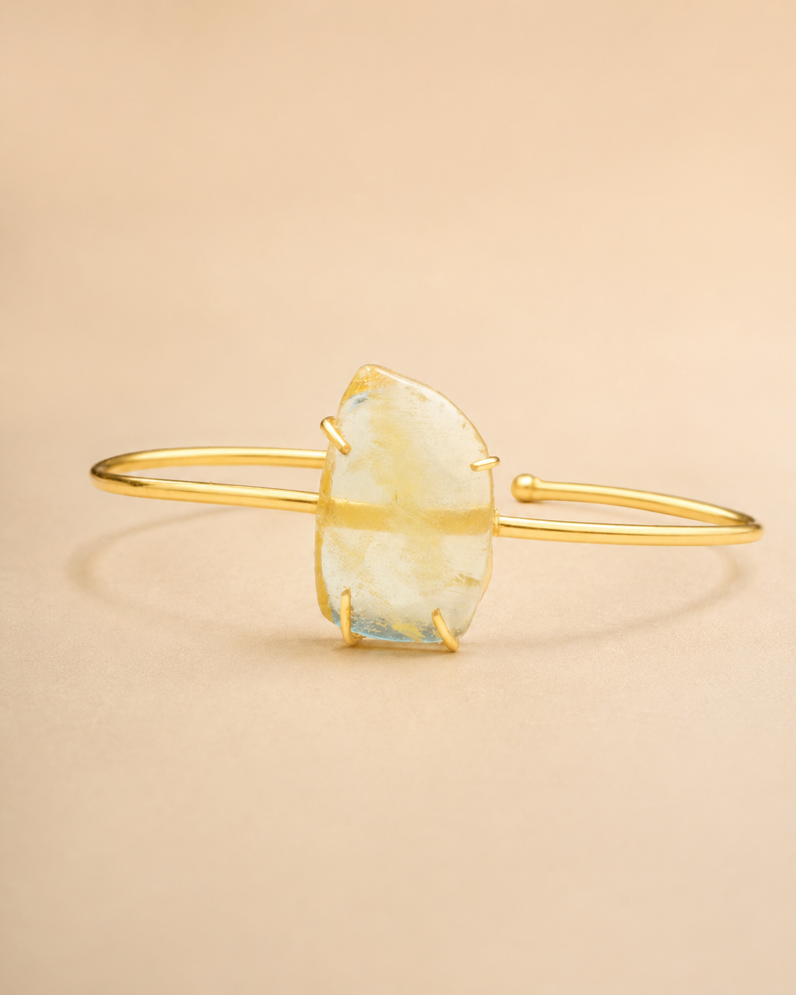 A gold bangle cuff bracelet featuring a large, irregularly shaped pale yellow and light blue raw stone, photographed on a solid beige background.