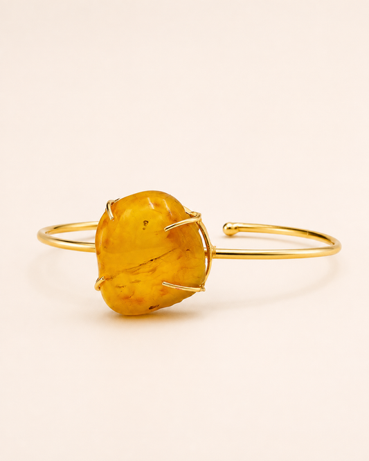 A gold-plated open cuff bangle bracelet featuring a large, organic-shaped yellow amber stone in a prong setting, shown on a warm off-white background.