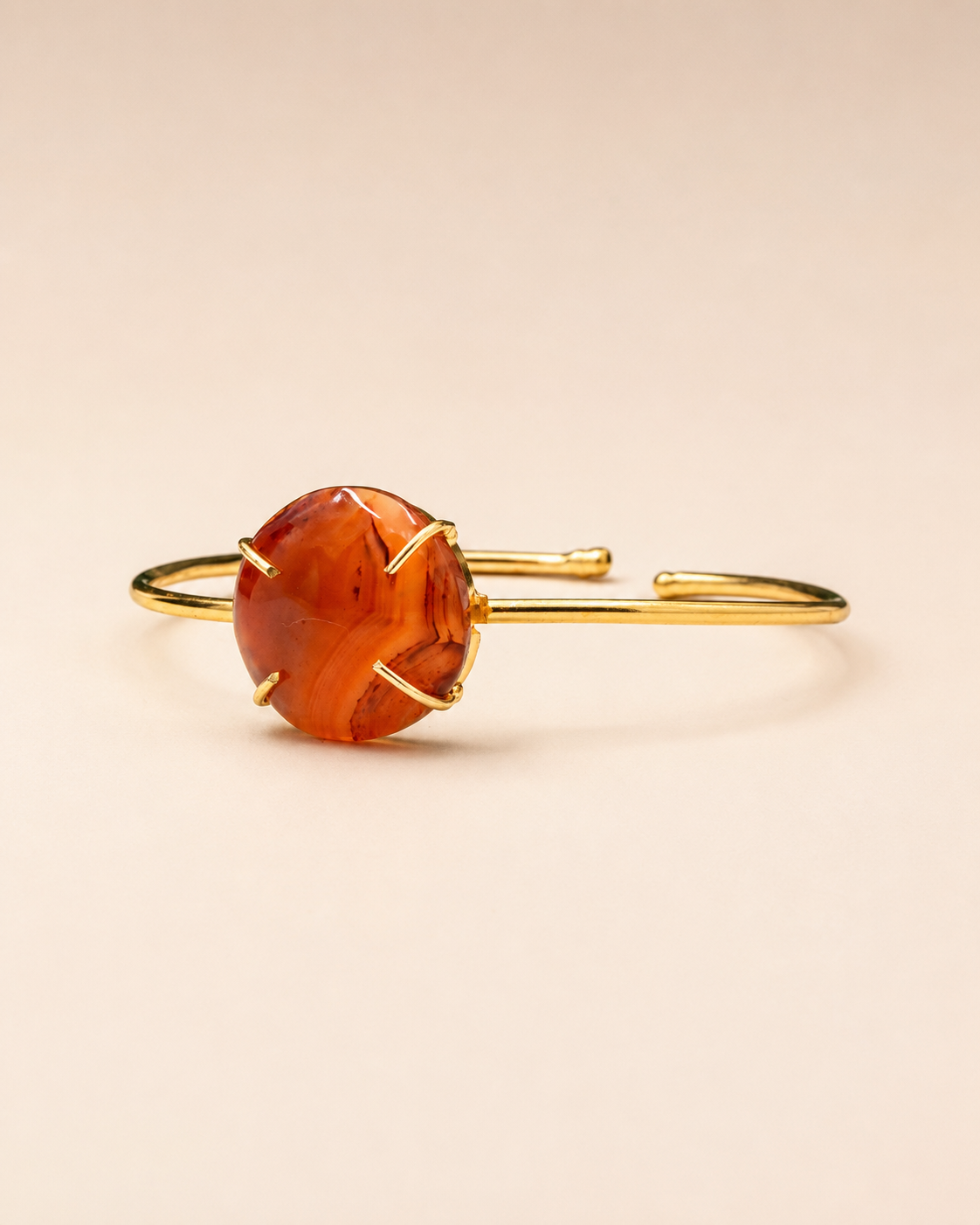 A gold-toned open cuff bracelet featuring a large, round, polished amber-orange cabochon gemstone in a prong setting, displayed on a solid light beige background.