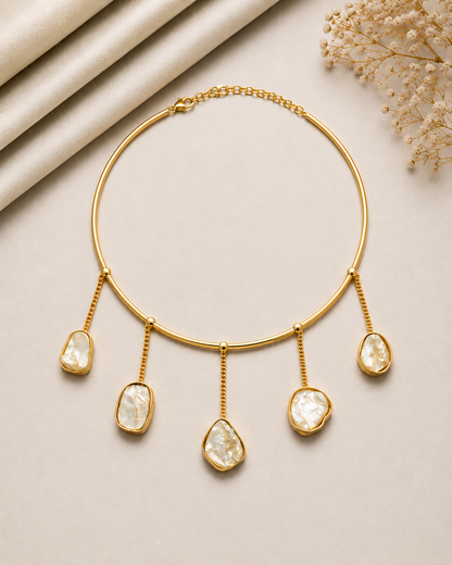 Gold-toned collar necklace with five irregular pear-shaped white stone drops, styled on a beige background with rolled satin fabric and dried flowers.