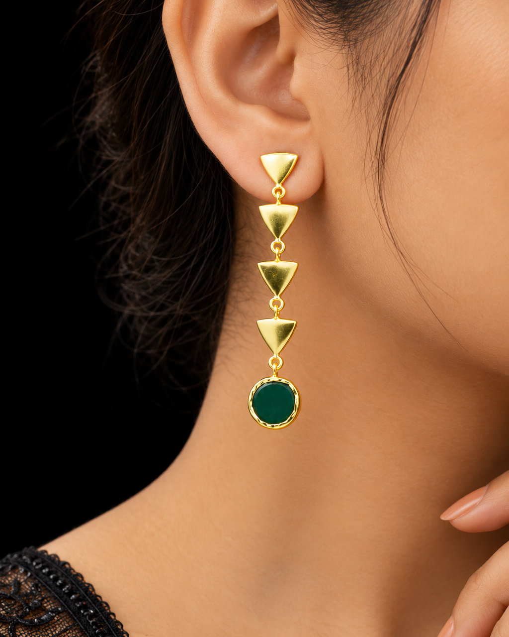 A close-up of a model's ear wearing a gold dangle earring with four stacked triangular gold links and a round dark green stone at the bottom.