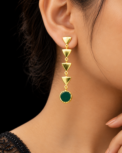 A close-up of a model's ear wearing a gold dangle earring with four stacked triangular gold links and a round dark green stone at the bottom.