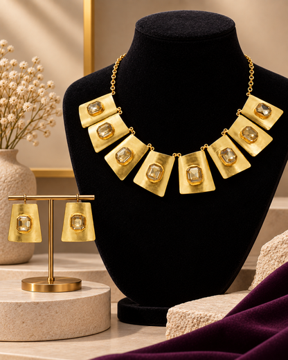 Gold statement necklace and matching drop earrings displayed on a black velvet mannequin bust and a gold stand, with a beige tabletop, dried flowers, and a draped purple fabric in the background.