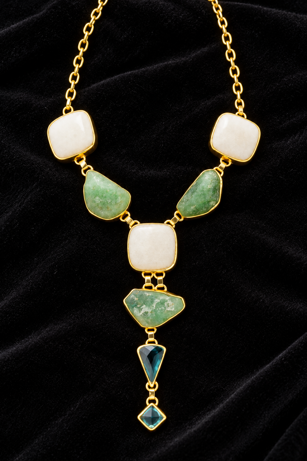 A gold-tone statement necklace featuring a central cream-colored stone, flanked by green marbled stones and a dangling green triangular charm, displayed on a black velvet background.