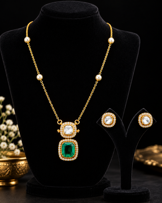 Gold necklace and matching stud earrings displayed on black velvet jewellery stands against a dark background. The necklace has a gold chain with pearl-like beads and a pendant featuring a large emerald-cut green stone and square clear stones set in gold.