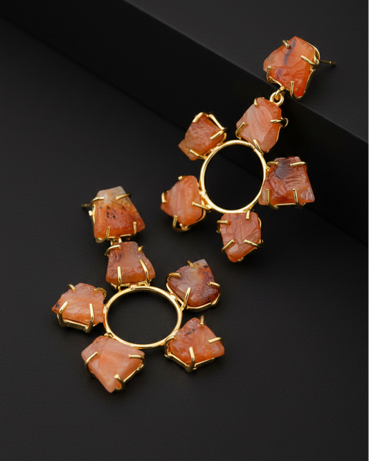 A pair of gold-tone statement earrings featuring irregular orange and peach agate stones arranged around a central gold hoop, displayed on a textured beige stone surface.