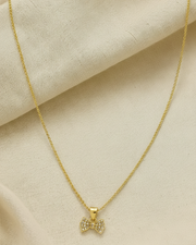A delicate gold chain necklace with a small bow-shaped pendant encrusted with clear stones, displayed on a draped beige fabric background.