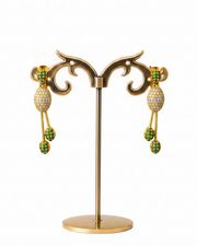 A pair of gold dangle earrings with green and white pavé details, displayed on a gold stand against a white background.