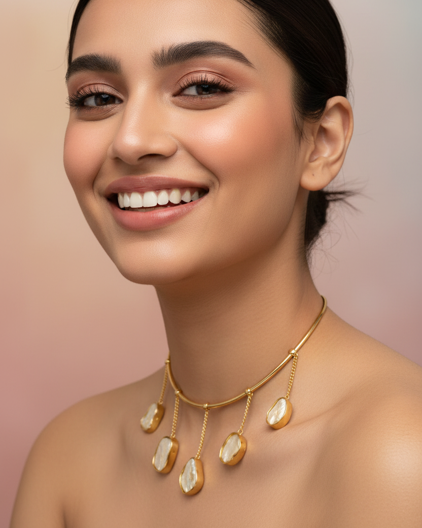 A close-up of a smiling woman wearing a gold necklace with irregular cream-colored stone pendants, set against a soft pink background.