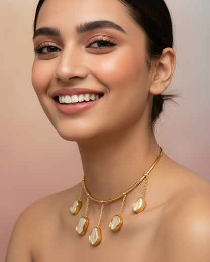 A close-up of a smiling woman wearing a gold necklace with irregular cream-colored stone pendants, set against a soft pink background.