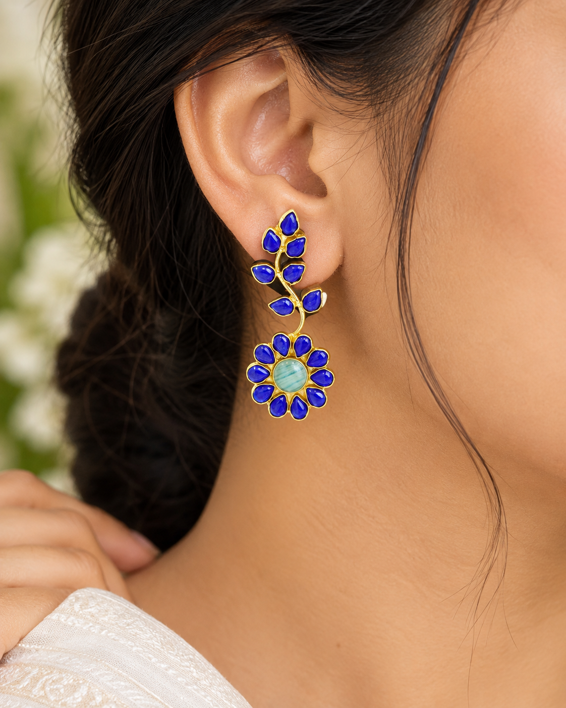 A close-up of a woman's ear wearing a gold dangle earring with deep blue stones and a central light blue-green stone.