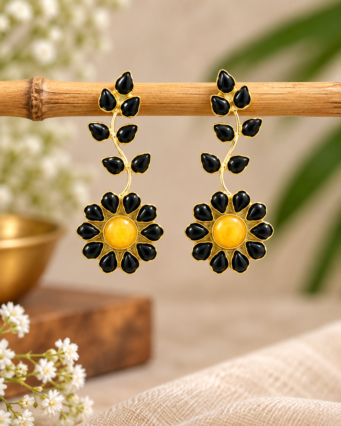 A pair of gold and black dangle earrings with a floral sunburst design and leaf motifs, hanging from a bamboo rod against a softly blurred neutral background.