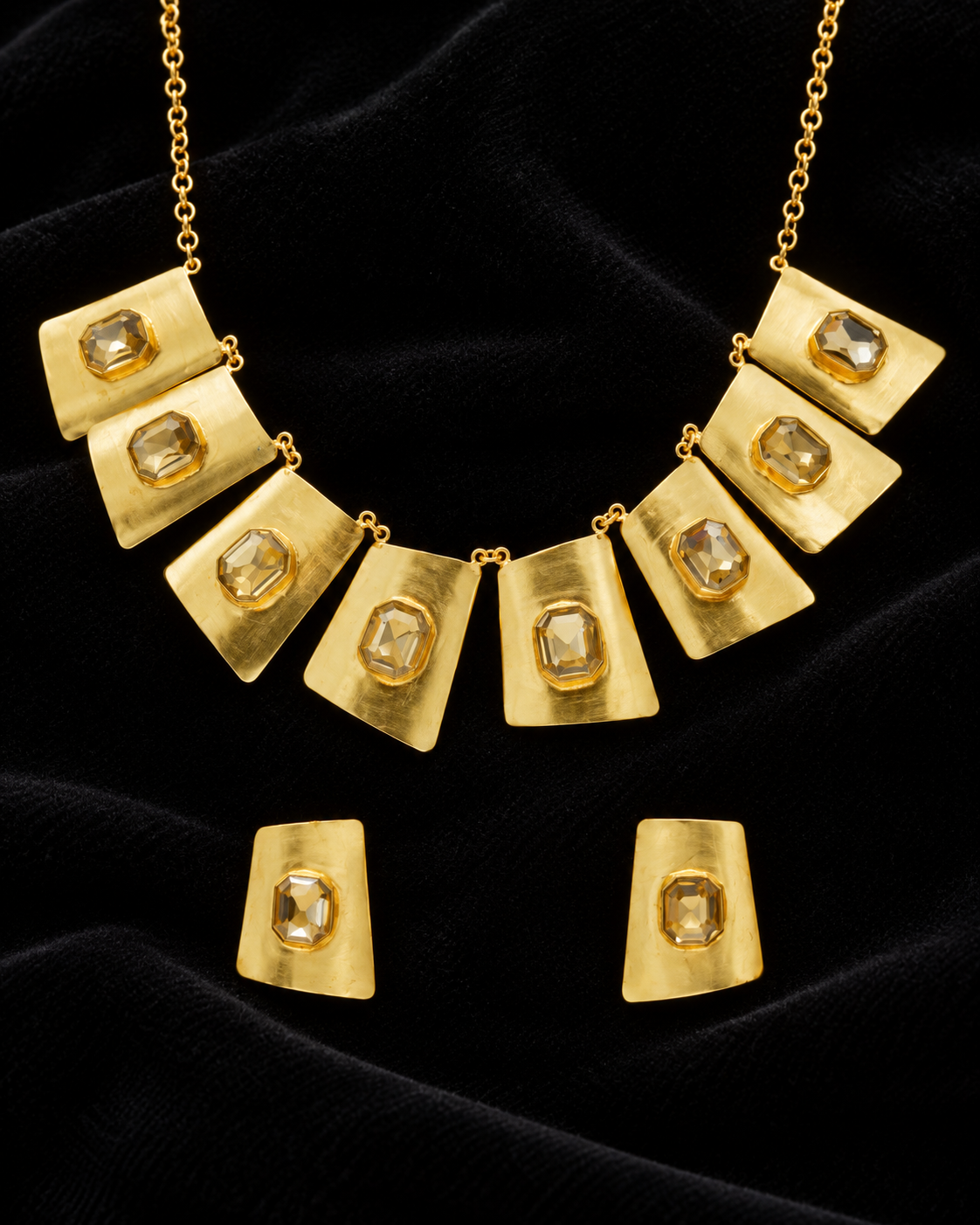 Gold statement necklace and matching earrings with a geometric, layered square design and clear faceted gemstones, displayed on a black velvet background.