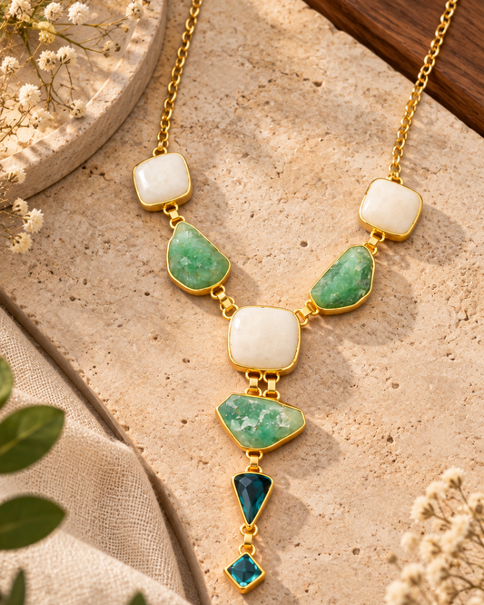 A gold-tone statement necklace with white and green stones, displayed on a textured beige stone surface with dried flowers and green leaves.