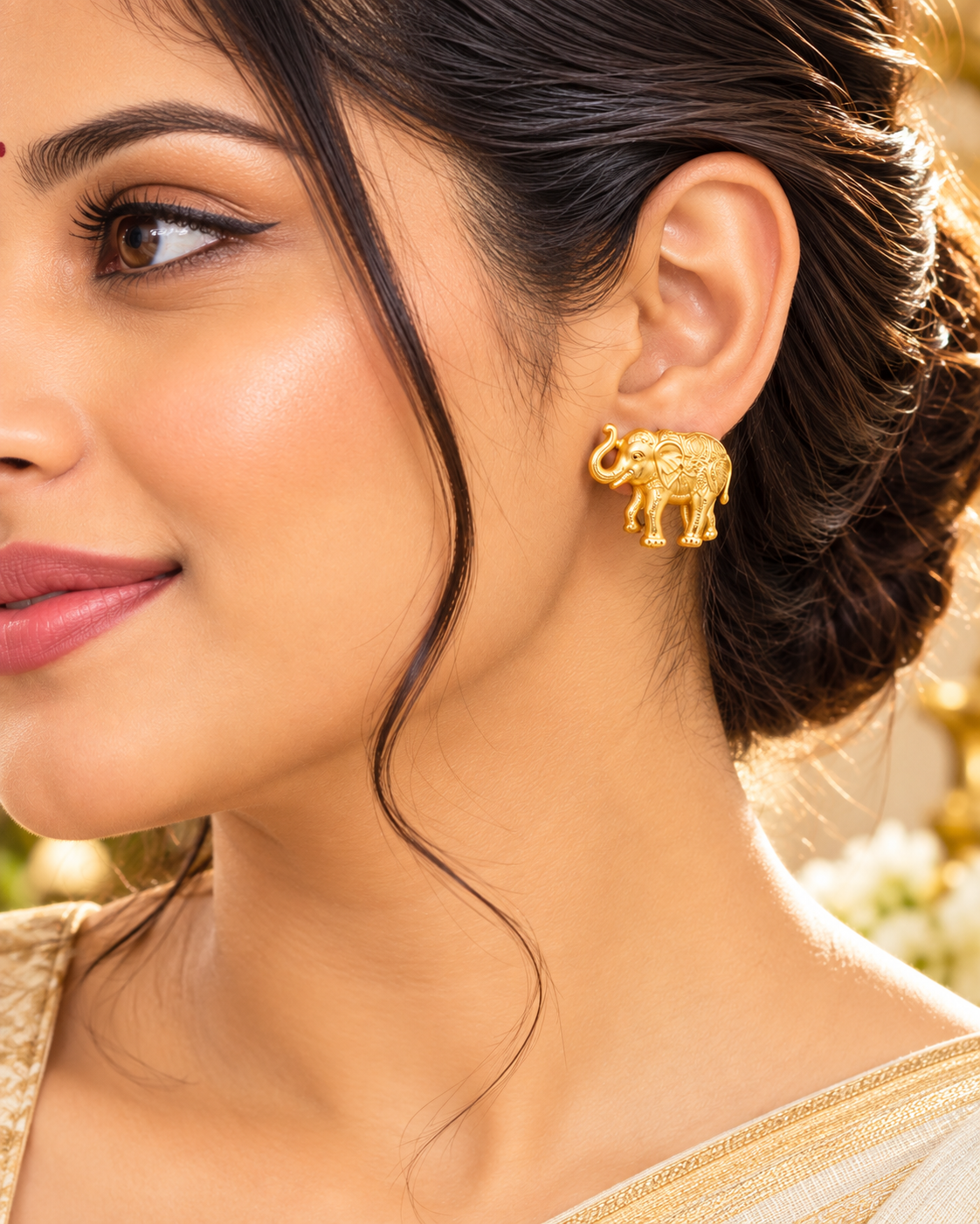 A close-up of a woman wearing a small gold elephant stud earring, with her ear, hair, and part of her clothing visible against a softly blurred warm background.