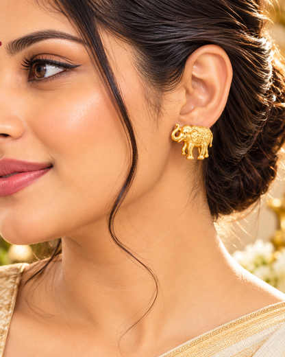 A close-up of a woman wearing a small gold elephant stud earring, with her ear, hair, and part of her clothing visible against a softly blurred warm background.
