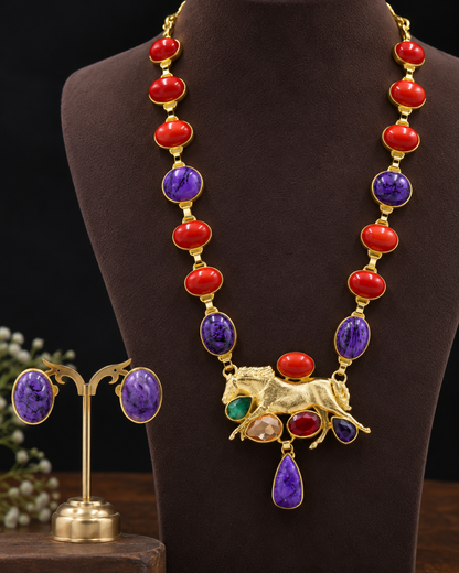 Gold-tone statement necklace with alternating red and purple oval and teardrop gemstones, plus a gold horse-shaped centerpiece, displayed on a dark brown jewelry bust. Matching stud earrings are shown on a small stand to the left.