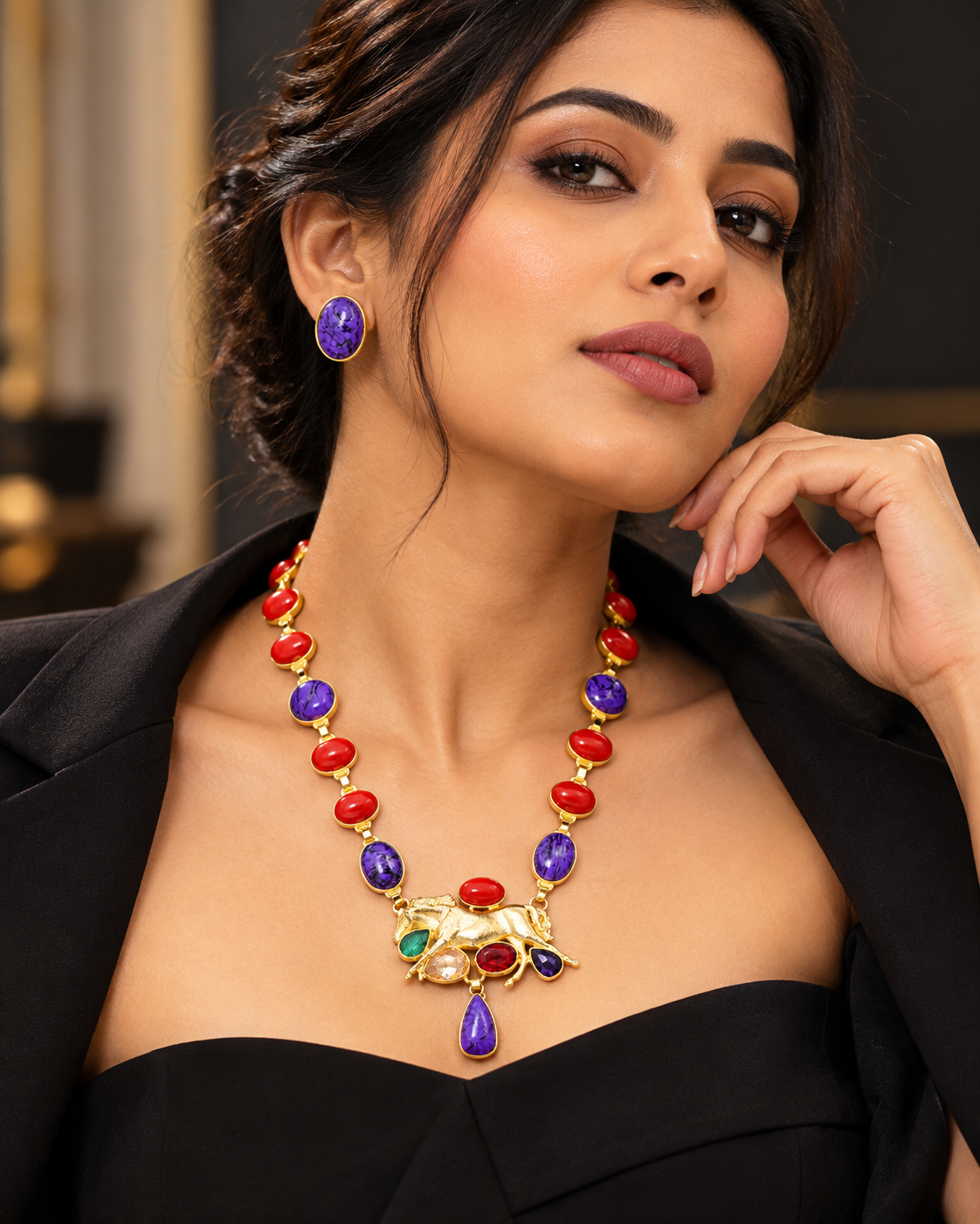 A woman wearing a gold-plated statement necklace and matching stud earring. The necklace features a detailed elephant pendant and a drop pendant, both decorated with red, purple, and green stones, and is accented by red and purple stones along the chain.