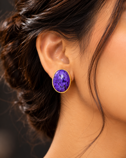 A close-up of a woman's ear wearing a gold stud earring with an oval purple stone featuring a carved face design.