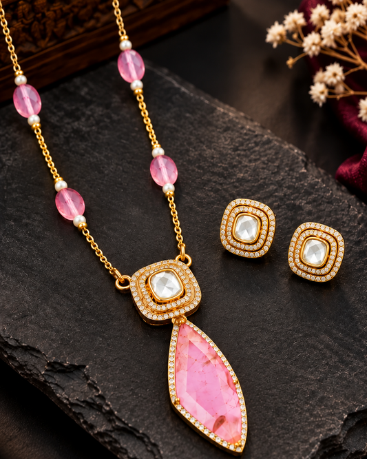 Gold-tone necklace and matching stud earrings with pink gemstone accents, displayed on a dark stone surface with decorative dried flowers and a wooden box in the background.