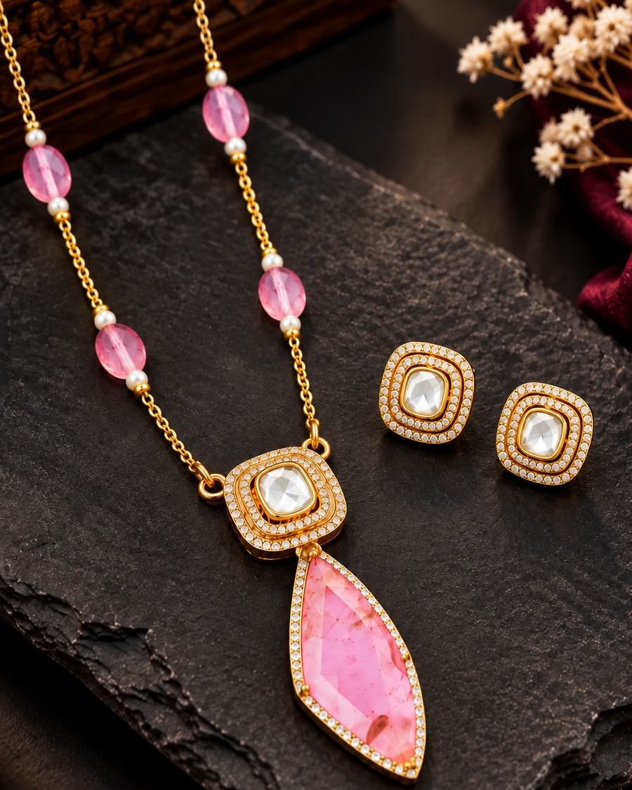 Gold-tone necklace and matching stud earrings with pink gemstone accents, displayed on a dark stone surface with decorative dried flowers and a wooden box in the background.