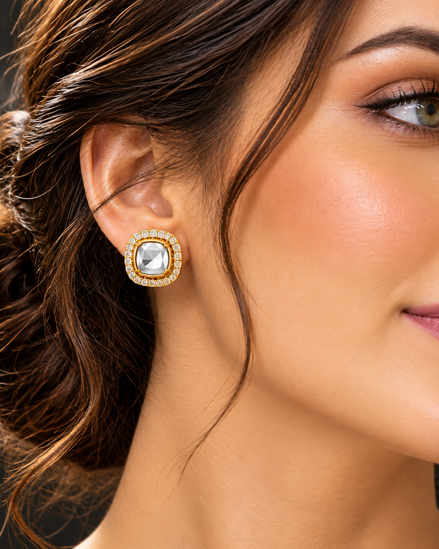 A close-up of a woman's ear wearing a square gold-toned earring with a clear central stone and a border of small clear stones.