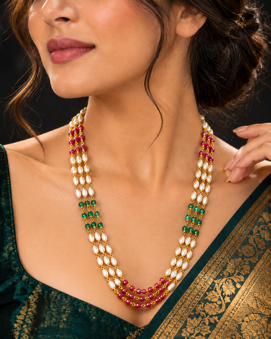 A close-up of a woman wearing a multi-strand traditional necklace with white pearl-like beads, gold-toned settings, green beads, and red beads, paired with a dark green saree.