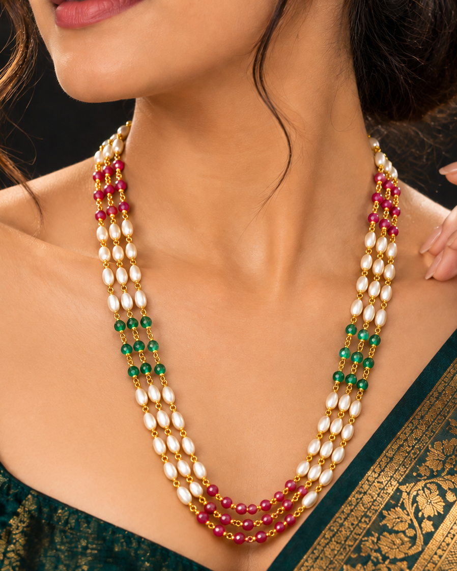 A close-up of a woman wearing a multi-strand traditional necklace with white pearl-like beads, gold-toned settings, green beads, and red beads, paired with a dark green saree.