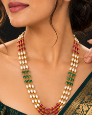 A close-up of a woman wearing a multi-strand traditional necklace with white pearl-like beads, gold-toned settings, green beads, and red beads, paired with a dark green saree.