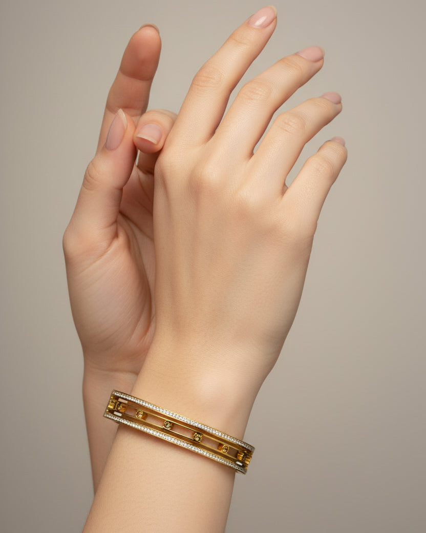A close-up of a person's hand and wrist wearing a double-strand gold bangle bracelet with pavé-set clear stones, set against a neutral warm gray background.