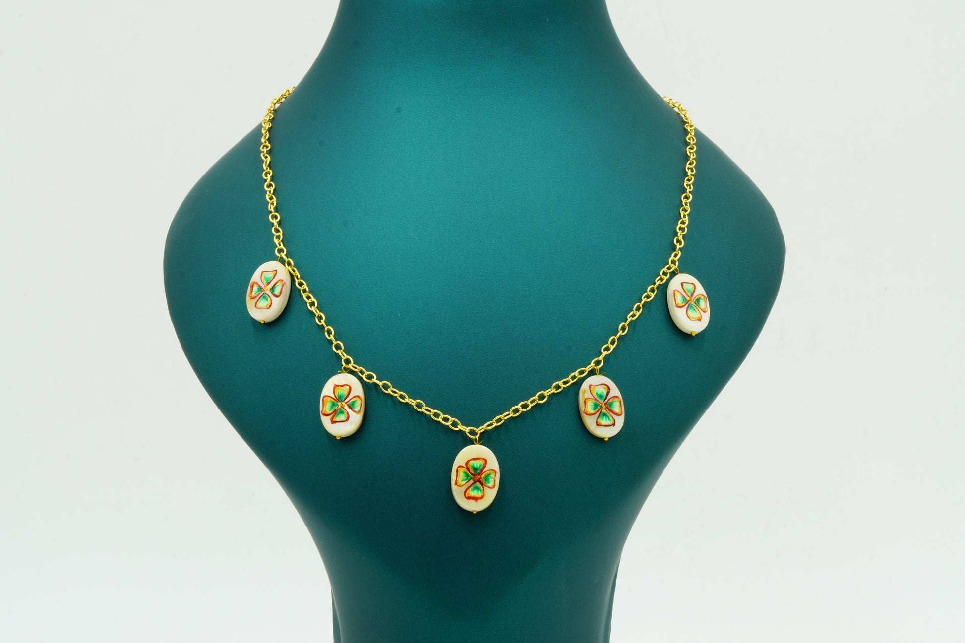 A gold chain necklace with five oval white enamel pendants featuring green clover motifs, displayed on a dark teal jewelry bust against a white background.