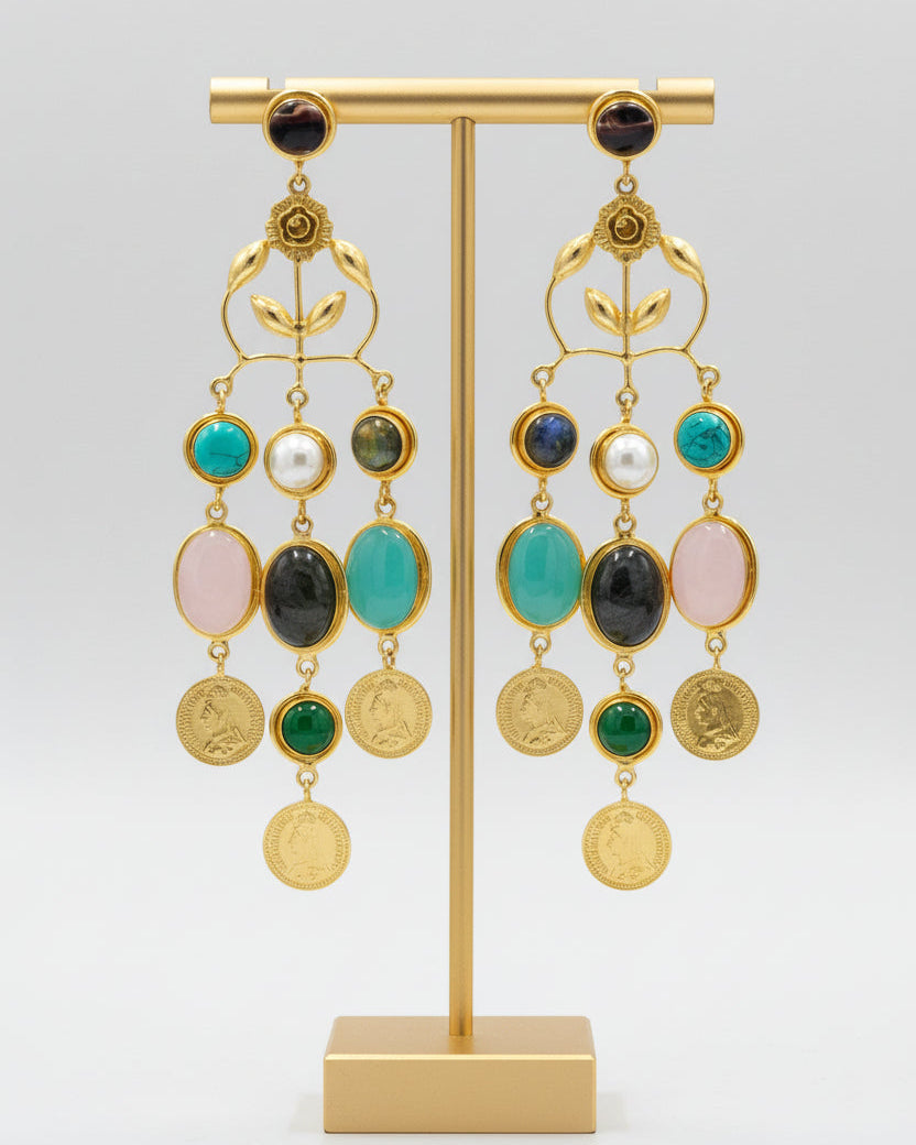 A pair of gold-toned statement dangle earrings displayed on a black velvet jewelry bust against a white background. The earrings feature multiple colorful gemstone drops including turquoise, white, pink, green, and dark gray, with round gold coin-like charms at the bottom.