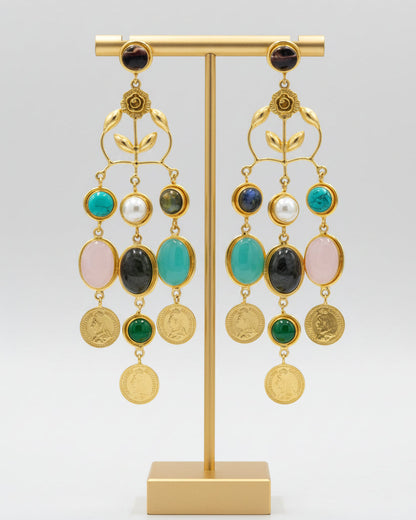 A pair of gold-toned statement dangle earrings displayed on a black velvet jewelry bust against a white background. The earrings feature multiple colorful gemstone drops including turquoise, white, pink, green, and dark gray, with round gold coin-like charms at the bottom.