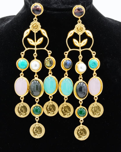 A pair of gold-toned statement dangle earrings displayed on a black velvet jewelry bust against a white background. The earrings feature multiple colorful gemstone drops including turquoise, white, pink, green, and dark gray, with round gold coin-like charms at the bottom.