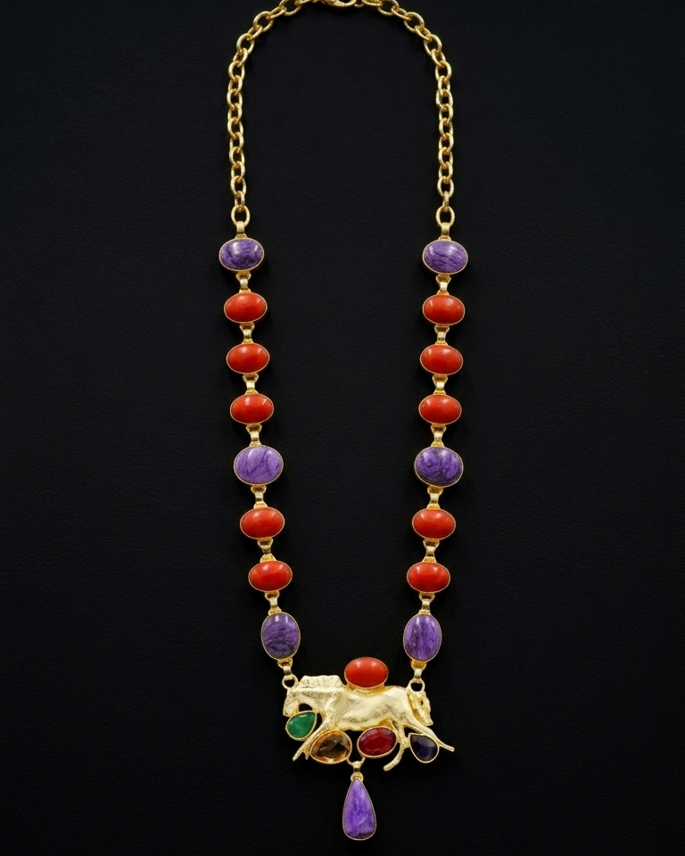Gold-tone jewelry set featuring a beaded necklace with red coral-like beads, purple and green stone accents, and a central animal motif with red and green stones, displayed above matching oval stud earrings on a black background.