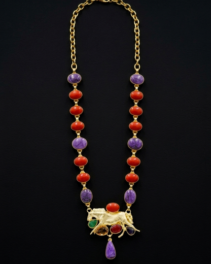 Gold-tone jewelry set featuring a beaded necklace with red coral-like beads, purple and green stone accents, and a central animal motif with red and green stones, displayed above matching oval stud earrings on a black background.